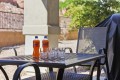 Outdoor patio scene with two Sunkist orange soda bottles and four empty glasses on a perforated metal table, surrounded by chairs.