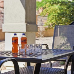Outdoor patio scene with two Sunkist orange soda bottles and four empty glasses on a perforated metal table, surrounded by chairs.