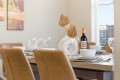 Dining table set for a meal with white dishes, multiple wine glasses, a bottle of red wine, and a white vase centerpiece with dried leaves.