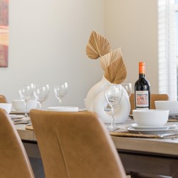 Dining table set for a meal with white dishes, multiple wine glasses, a bottle of red wine, and a white vase centerpiece with dried leaves.