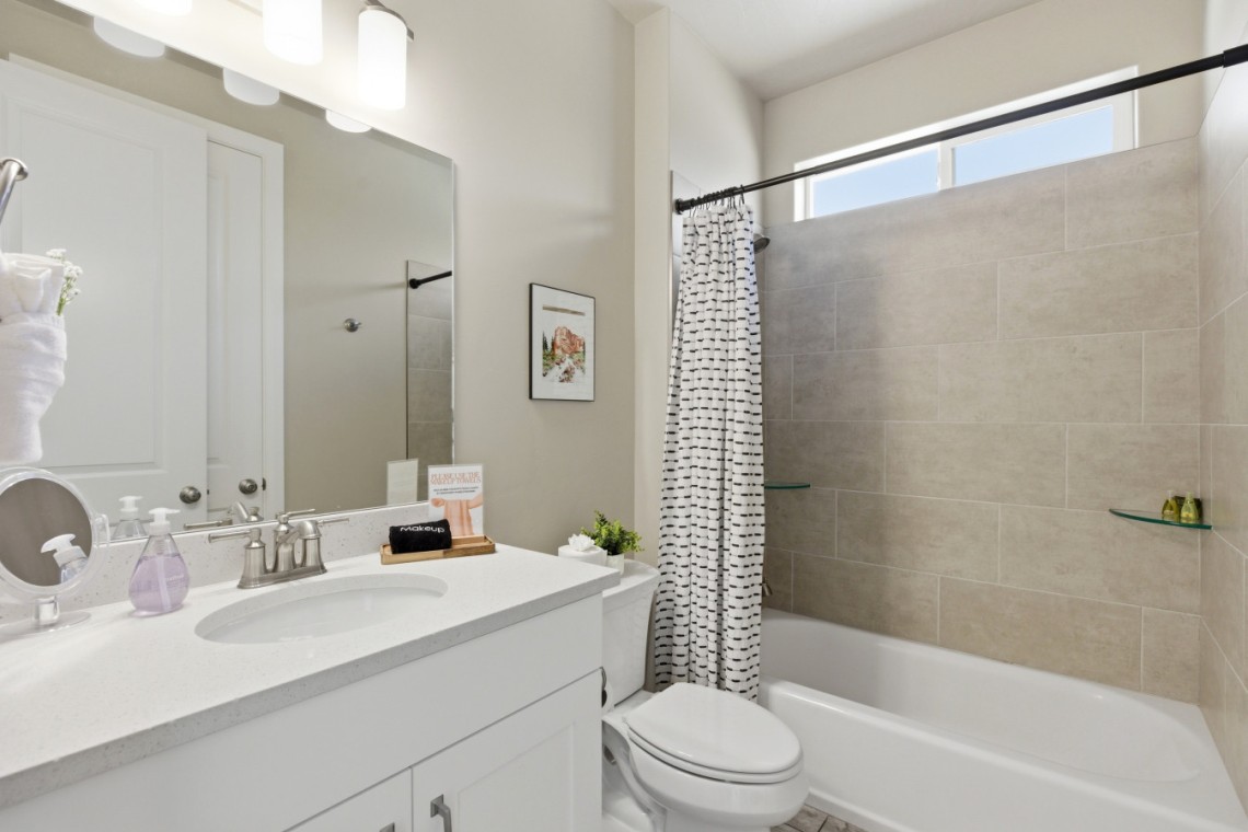 Modern bathroom with white vanity, oval sink, and large mirror; beige tiled tub/shower and striped curtain nearby.
