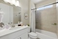 Modern bathroom with white vanity, oval sink, and large mirror; beige tiled tub/shower and striped curtain nearby.