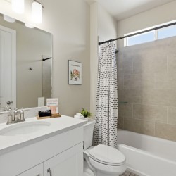 Modern bathroom with white vanity, oval sink, and large mirror; beige tiled tub/shower and striped curtain nearby.