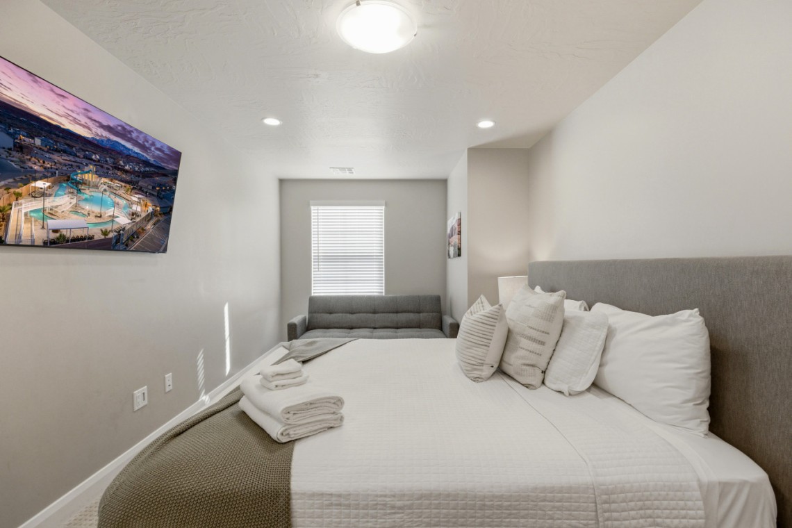 Cozy modern bedroom with a white quilted bed, gray headboard and pillows, and a gray sofa at the far end; wall-mounted TV on the left wall.