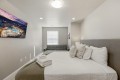 Cozy modern bedroom with a white quilted bed, gray headboard and pillows, and a gray sofa at the far end; wall-mounted TV on the left wall.