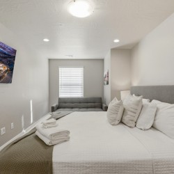 Cozy modern bedroom with a white quilted bed, gray headboard and pillows, and a gray sofa at the far end; wall-mounted TV on the left wall.