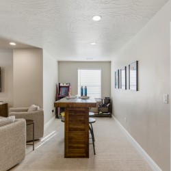Basement recreation room with a wooden bar, two beige armchairs, and a wall-mounted TV showing an outdoor scene, plus framed photos on the right wall.