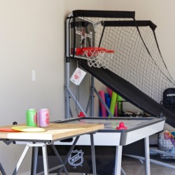 Indoor game room with a basketball arcade game and an air hockey table against a beige wall; two soda cans sit on a wooden table in the foreground.