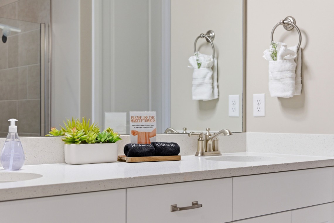 White bathroom counter with a planter of succulents, a small sign about makeup towels, and a tray with black rolled towels beside a brushed-metal faucet.