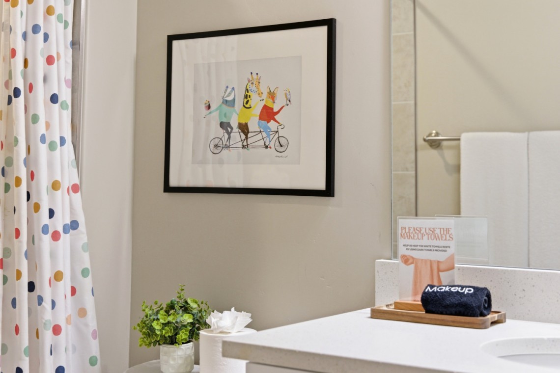 Framed colorful animal illustration on a gray bathroom wall, with a polka-dot shower curtain on the left and a white countertop with decor on the right.