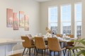 Dining room with a wooden table set for six, beige chairs, wine bottle centerpiece, and large windows with blinds.