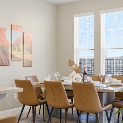 Dining room with a wooden table set for six, beige chairs, wine bottle centerpiece, and large windows with blinds.