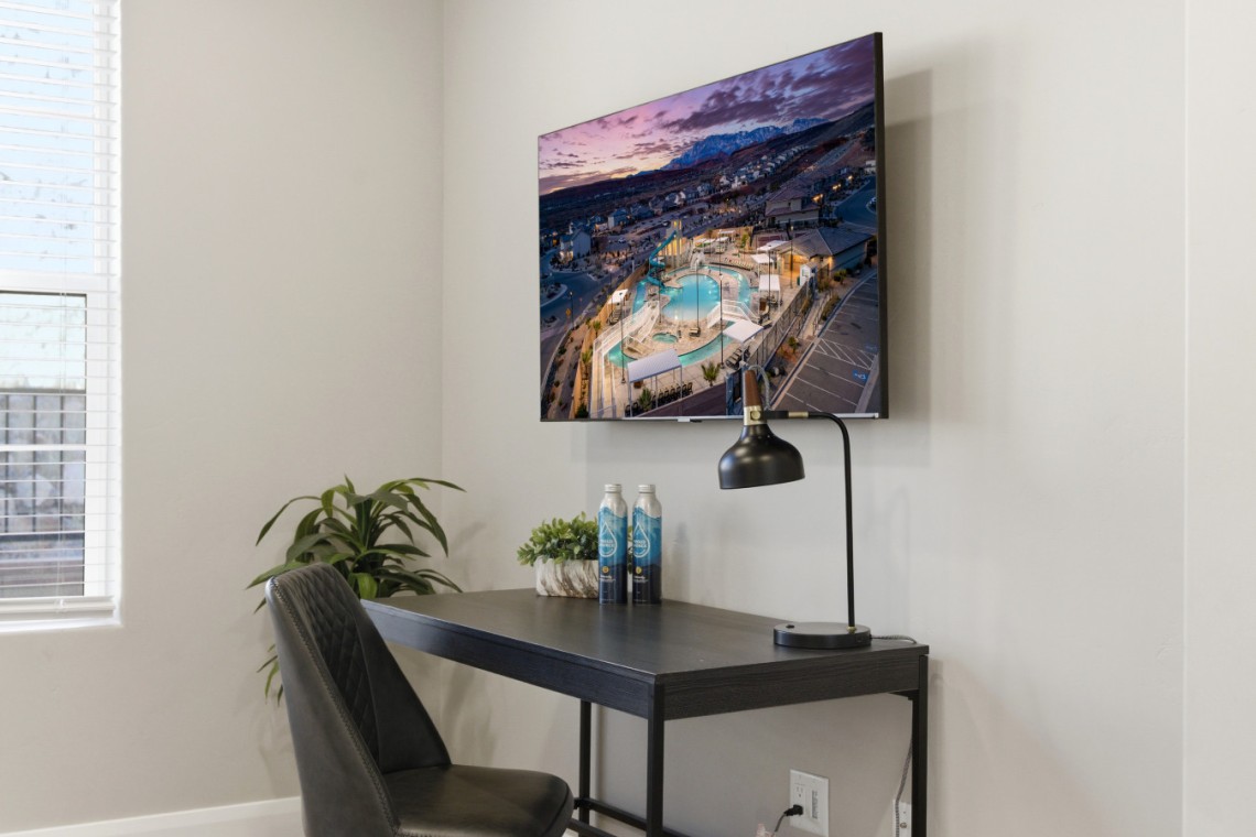 Desk setup with a wall-mounted TV showing an aerial pool scene, a black arc lamp, two water bottles, and a small plant on a dark desk.