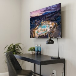 Desk setup with a wall-mounted TV showing an aerial pool scene, a black arc lamp, two water bottles, and a small plant on a dark desk.