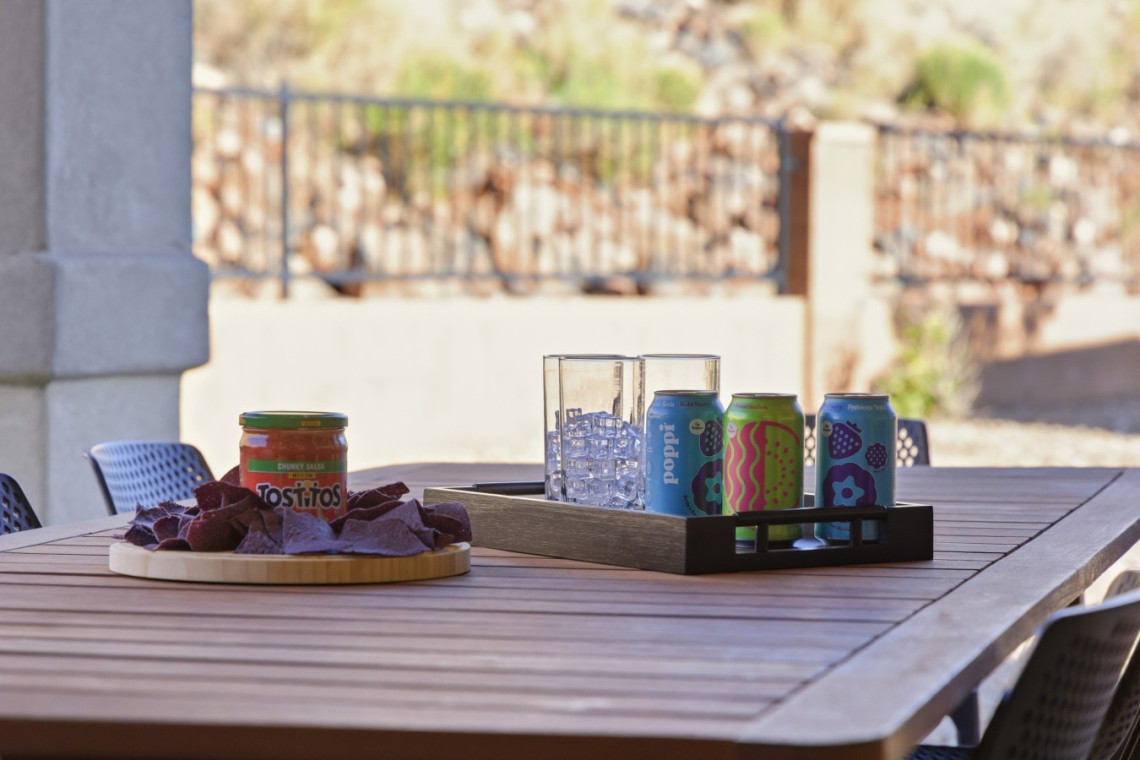 Outdoor patio table with a tray of colorful soda cans and a glass ice bucket, plus a jar of salsa and chips on a round board (snack setup).