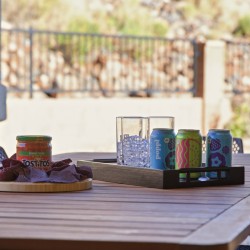 Outdoor patio table with a tray of colorful soda cans and a glass ice bucket, plus a jar of salsa and chips on a round board (snack setup).