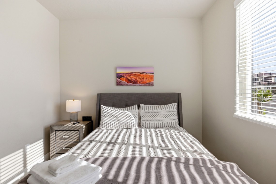 Minimalist bedroom with a gray upholstered bed, two striped pillows, a patterned duvet, and sunlit window blinds casting stripes on the bed