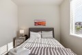 Minimalist bedroom with a gray upholstered bed, two striped pillows, a patterned duvet, and sunlit window blinds casting stripes on the bed