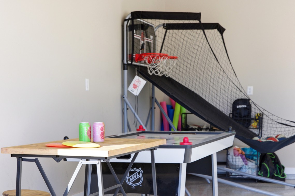 Indoor home game room with an air hockey table enclosed by a black net, a basketball hoop mounted on a backboard behind it, and a wooden tabletop with two soda cans in the foreground.