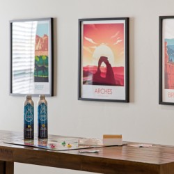 Wooden table with two blue bottled drinks and scattered board game pieces, against a wall with three framed travel posters (Reef, Arches, Bryce Canyon).