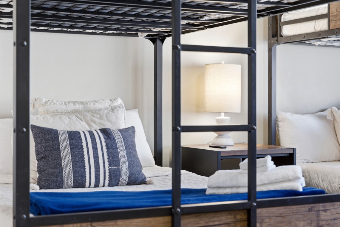 Metal bunk bed frame with white bedding and a blue striped pillow; a nightstand lamp sits between the beds, with folded towels at the foot of the bed.
