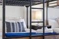 Metal bunk bed frame with white bedding and a blue striped pillow; a nightstand lamp sits between the beds, with folded towels at the foot of the bed.