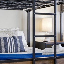 Metal bunk bed frame with white bedding and a blue striped pillow; a nightstand lamp sits between the beds, with folded towels at the foot of the bed.