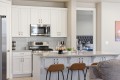 Bright white kitchen with stainless steel stove and microwave, glass subway tile backsplash, and island with three brown stools; doorway to a bedroom in view.