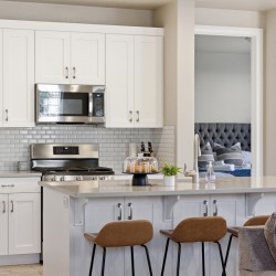 Bright white kitchen with stainless steel stove and microwave, glass subway tile backsplash, and island with three brown stools; doorway to a bedroom in view.