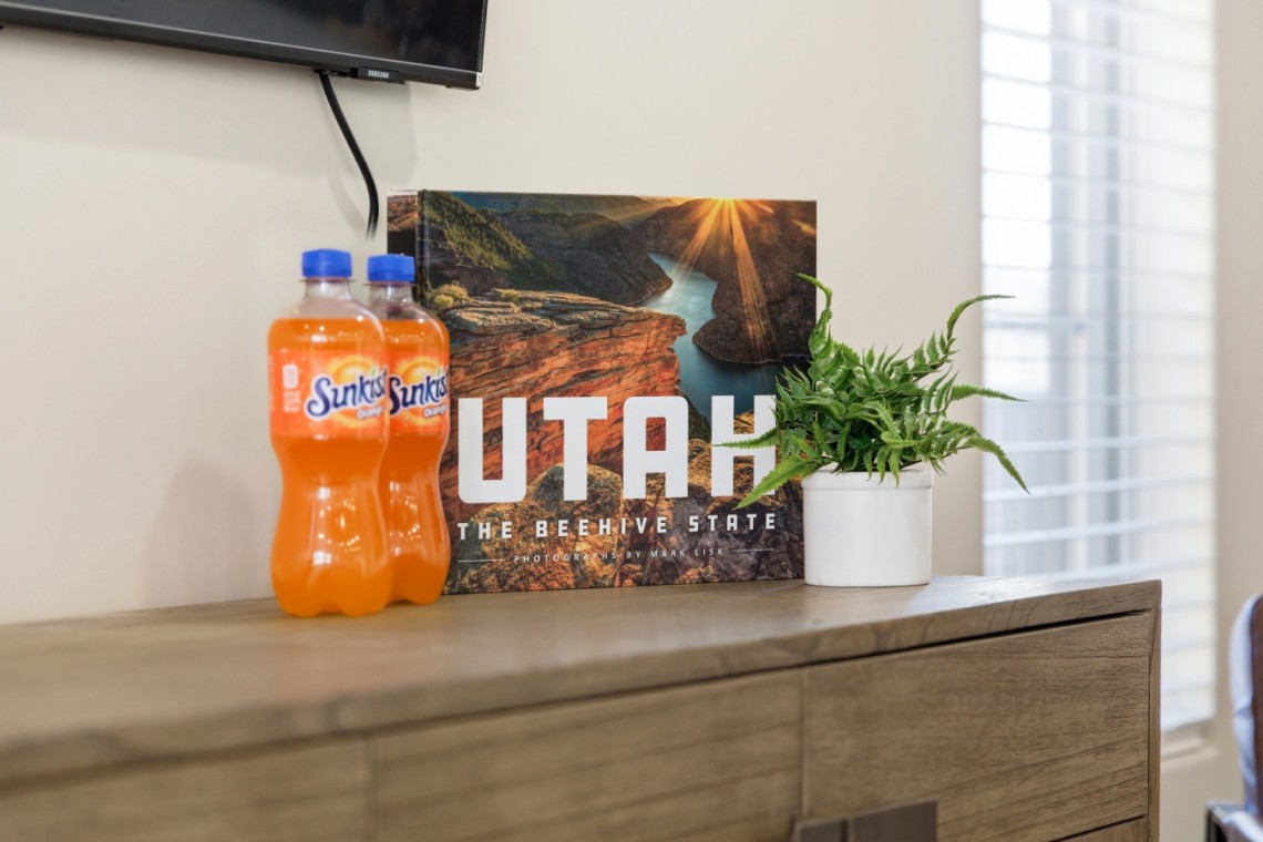 Two Sunkist orange soda bottles on a wooden dresser beside a Utah travel poster and a potted fern.