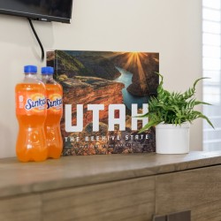 Two Sunkist orange soda bottles on a wooden dresser beside a Utah travel poster and a potted fern.