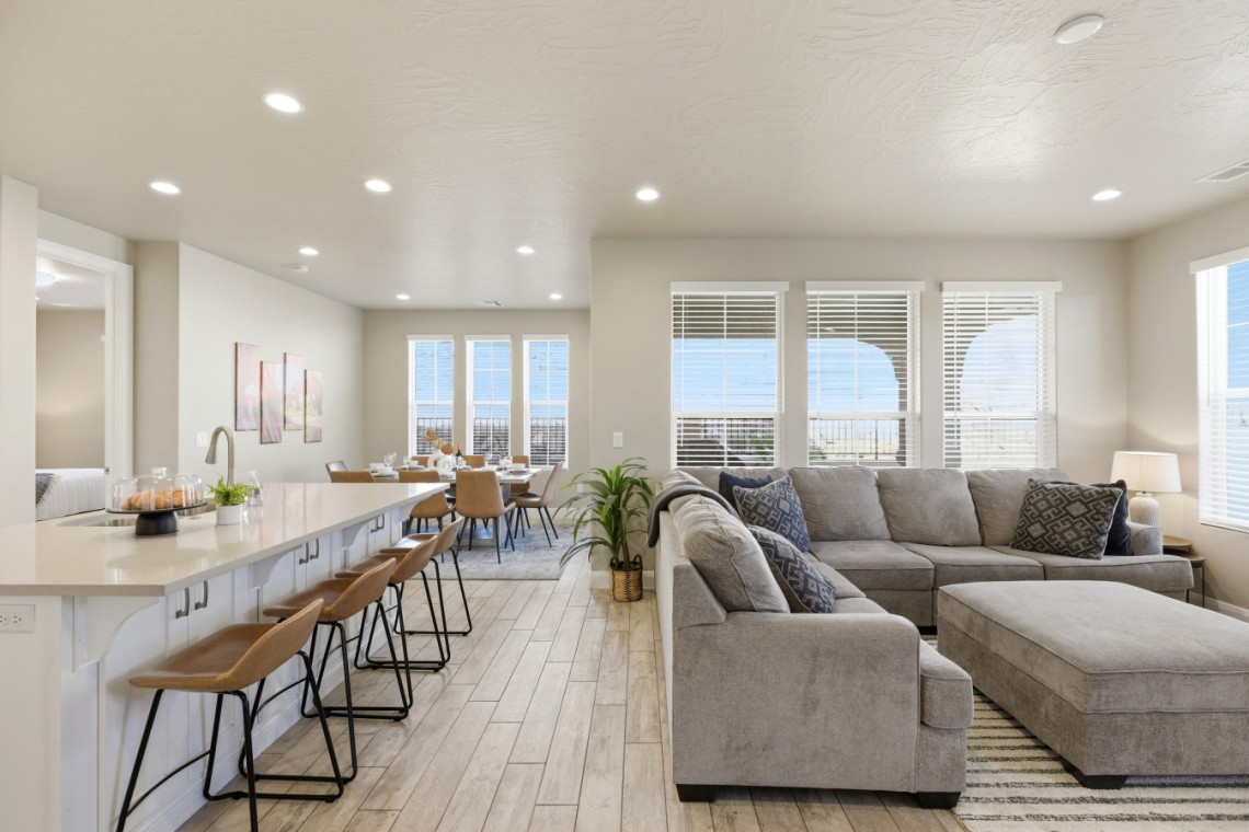 Bright open-concept kitchen and living area with a white island, brown bar stools, and a gray sectional by large windows with blinds.