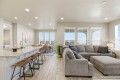 Bright open-concept kitchen and living area with a white island, brown bar stools, and a gray sectional by large windows with blinds.