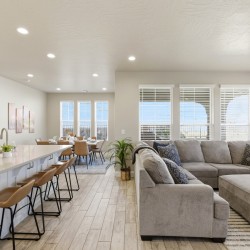 Bright open-concept kitchen and living area with a white island, brown bar stools, and a gray sectional by large windows with blinds.