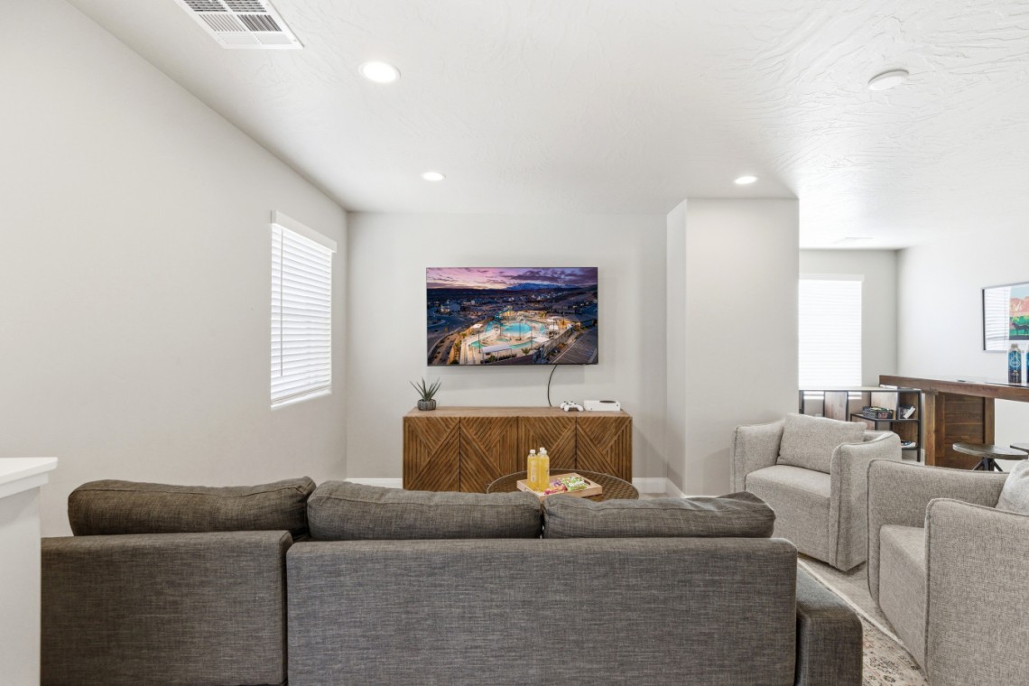 Neutral living room with gray sectional facing a wall-mounted flat-screen TV above a geometric wooden cabinet, plant, and game console.
