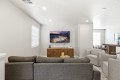 Neutral living room with gray sectional facing a wall-mounted flat-screen TV above a geometric wooden cabinet, plant, and game console.