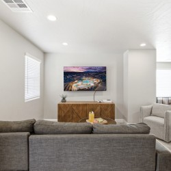 Neutral living room with gray sectional facing a wall-mounted flat-screen TV above a geometric wooden cabinet, plant, and game console.