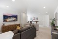 Living room with a wall-mounted TV above a wooden cabinet, gray sofa, beige armchairs, and a bar area in the background.