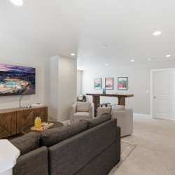 Living room with a wall-mounted TV above a wooden cabinet, gray sofa, beige armchairs, and a bar area in the background.