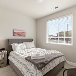 Gray upholstered bed with patterned bedding, gray throw, and folded towels on a nightstand in a bright bedroom.