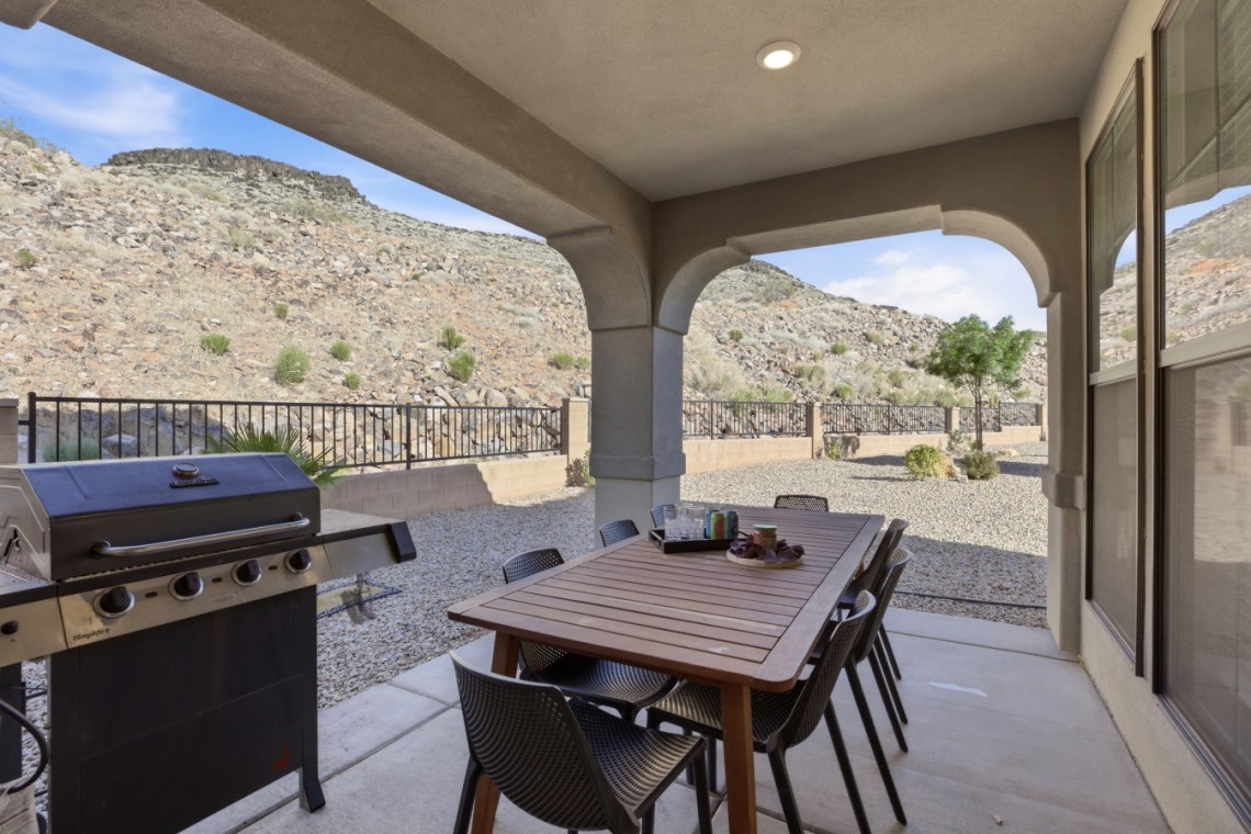 Covered patio with a wooden dining table and eight chairs, a gas grill on the left, and desert hills in the background.