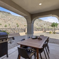 Covered patio with a wooden dining table and eight chairs, a gas grill on the left, and desert hills in the background.