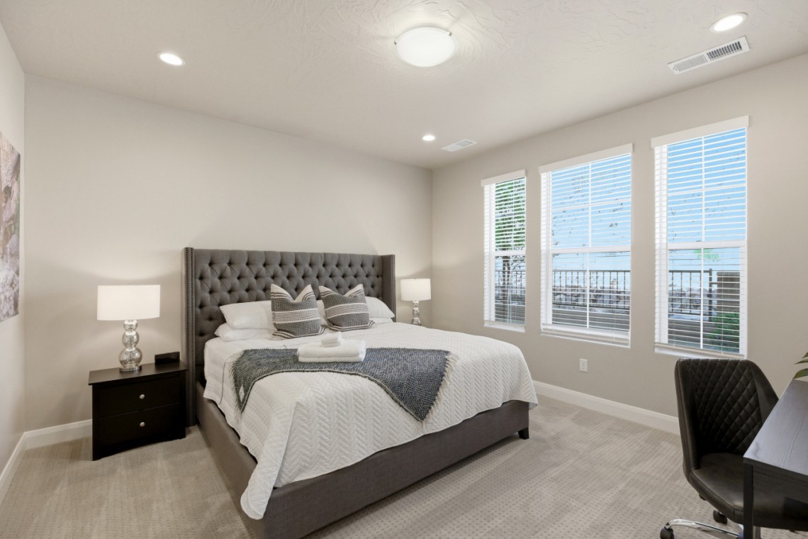 Modern bedroom with a tufted gray bed, white bedding, two glass-lamp nightstands, and three windows letting in light.