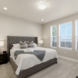 Modern bedroom with a tufted gray bed, white bedding, two glass-lamp nightstands, and three windows letting in light.