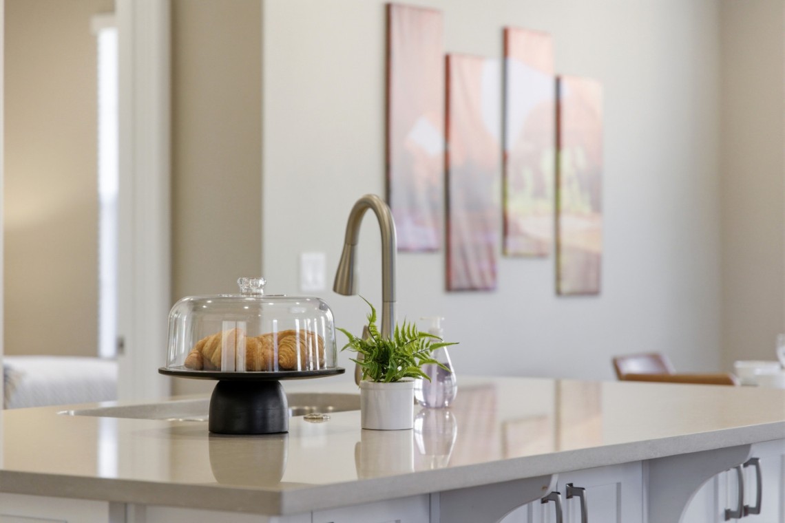 Croissants under a glass cake dome on a white kitchen island with a small potted plant nearby and a modern faucet in the background.