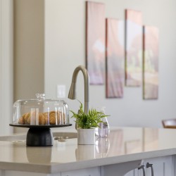 Croissants under a glass cake dome on a white kitchen island with a small potted plant nearby and a modern faucet in the background.