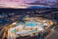 Aerial view of a community pool area with a twisting slide, lounge chairs, and shaded seating surrounded by houses at dusk, with mountains in the background.