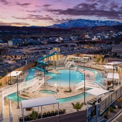Aerial view of a community pool area with a twisting slide, lounge chairs, and shaded seating surrounded by houses at dusk, with mountains in the background.