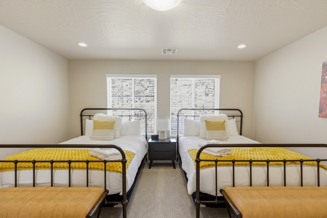 Twin bedroom with two metal-framed beds, white linens, and yellow knit throws, centered by a shared nightstand and lamp.