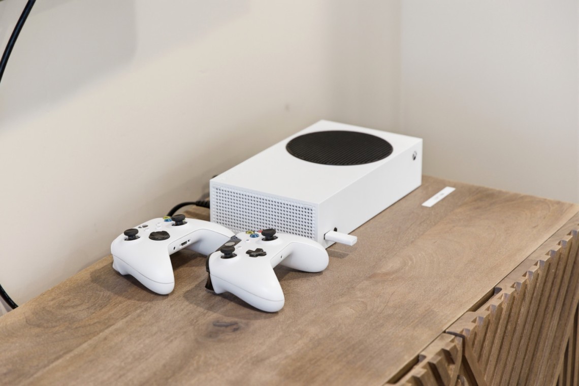 White Xbox Series S with two white controllers resting on a wooden table, plugged in against a neutral wall.